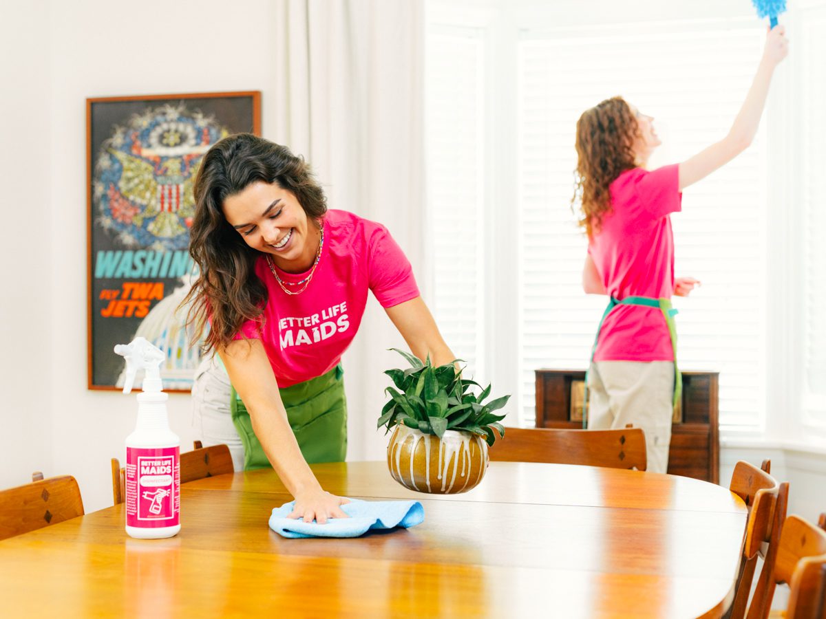 Better Life Maids professional house cleaning team providing top quality house cleaning in Webster Groves, MO. In this picture two Better Life Maids cleaning professionals can be seen house cleaning in Webster Groves. One professional cleaning technician is dusting a mid century modern dining room table and you can see she is a highly trained cleaning professional. She is taking care of her Webster Groves house cleaning customers detailed needs. The other cleaning professional in the background is high dusting the windows and blinds of a high end Webster Groves home.