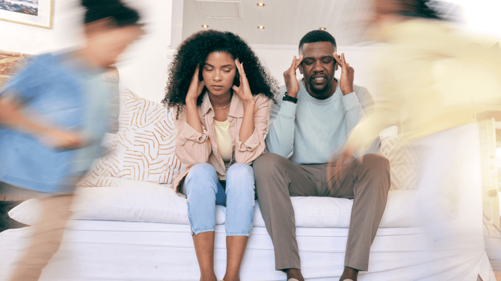 Parents sitting together looking mentally exhausted as children run around the living room at home.