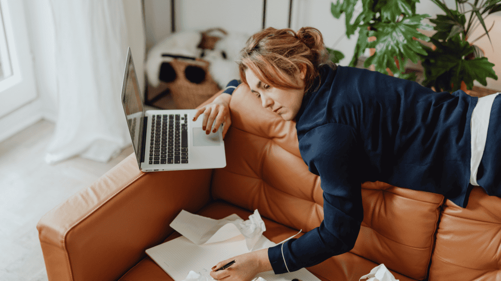 Woman leaning over a couch using a laptop while writing notes, looking mentally exhausted at home.